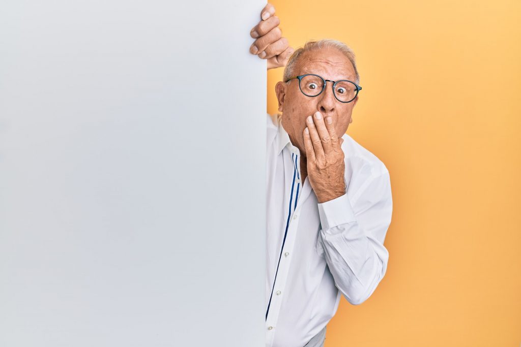 A grey-haired man in glasses peering around a corner with his hand to his mouth in a look of surprise