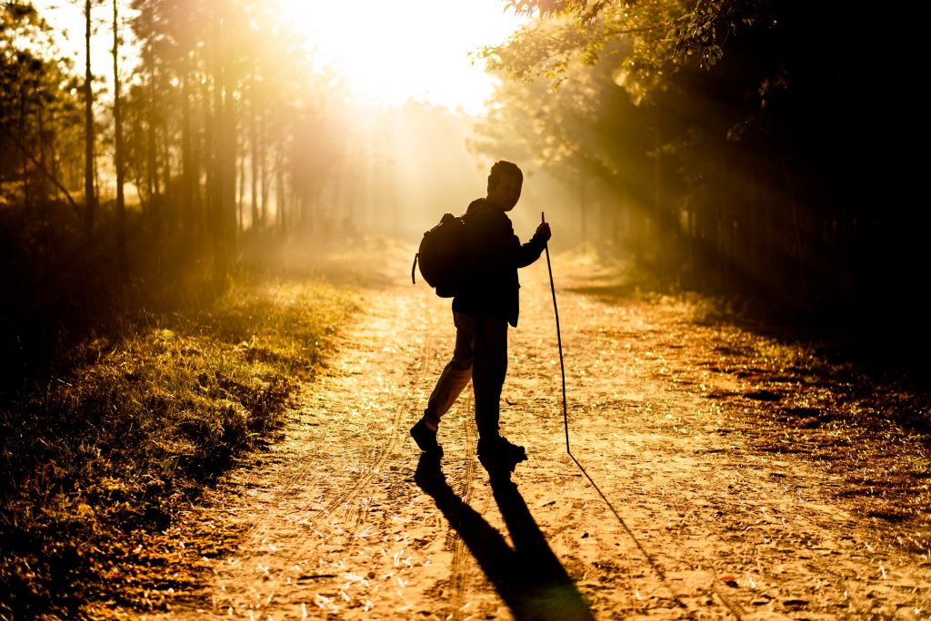 Man walking into the sunrise with a backpack and walking stick.